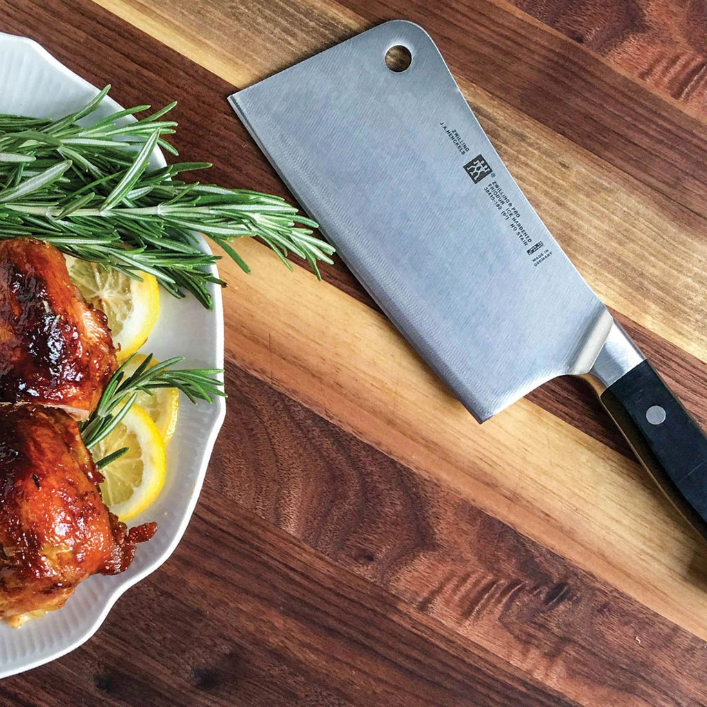 Meat cleaver with stainless blade and black handle on wooden board beside roast and herbs