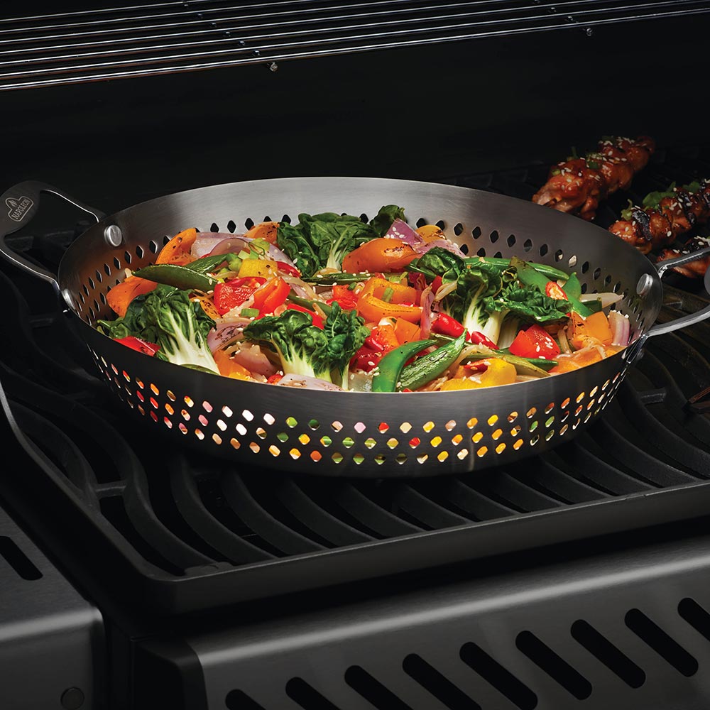 Perforated stainless steel grilling wok filled with vegetables on a grill