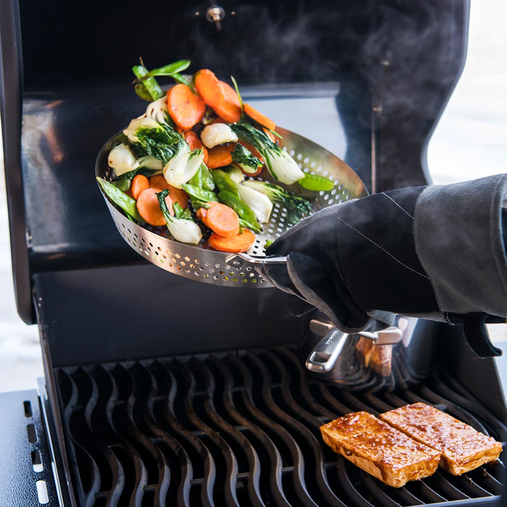Perforated grilling wok with tongs serving vegetables over a grill grate