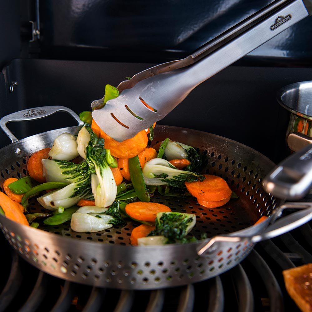 Perforated stainless steel wok holding sauteing vegetables over an open grill