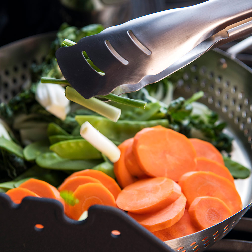 Stainless steel grilling wok with sliced carrots and greens being lifted by spatula
