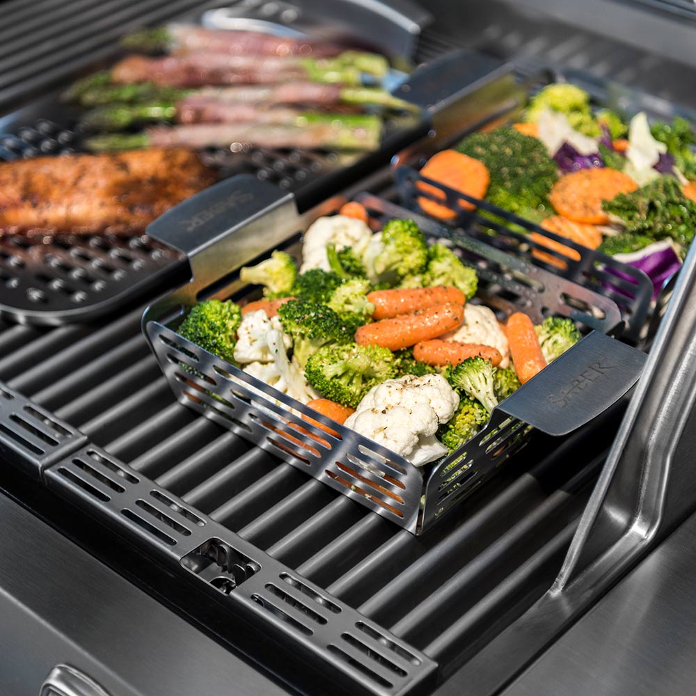 Stainless steel veggie basket holding vegetables and herbs on grill grates, Saber handle