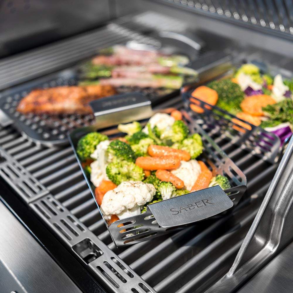 Stainless steel veggie basket with mixed vegetables and herbs on grill grates