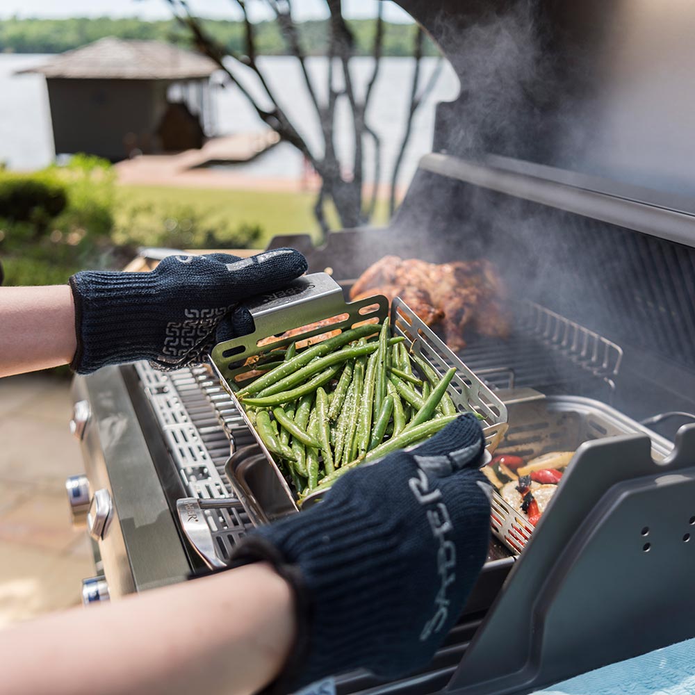 Hand placing green vegetables into stainless steel veggie basket on outdoor grill