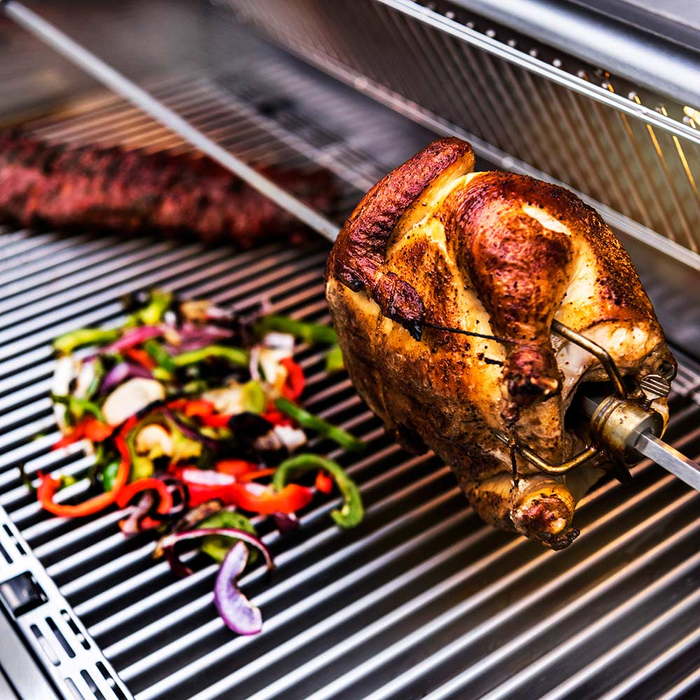 Rotisserie chicken on spit rod beside vegetable tray on grill grates