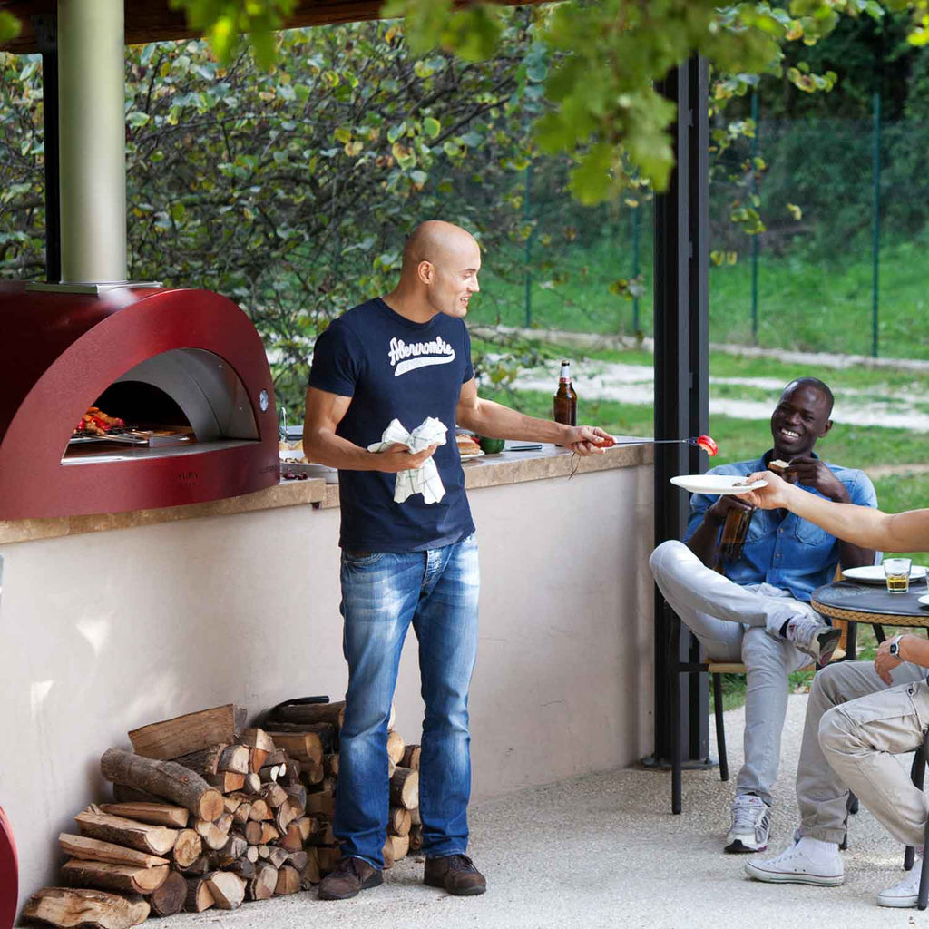 Dark red countertop wood-fired pizza oven with front arch and wood-log storage area
