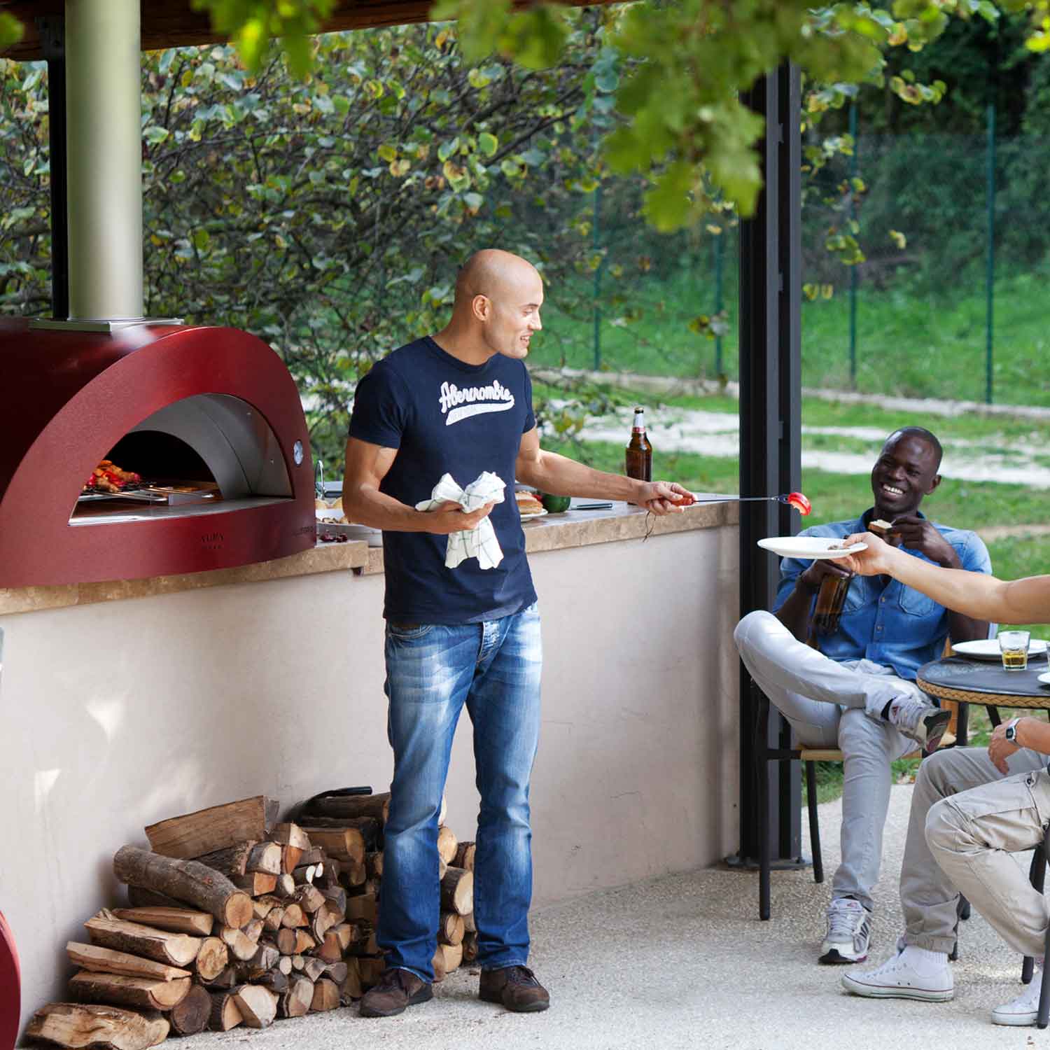 Dark red countertop wood-fired pizza oven with front arch and wood-log storage area