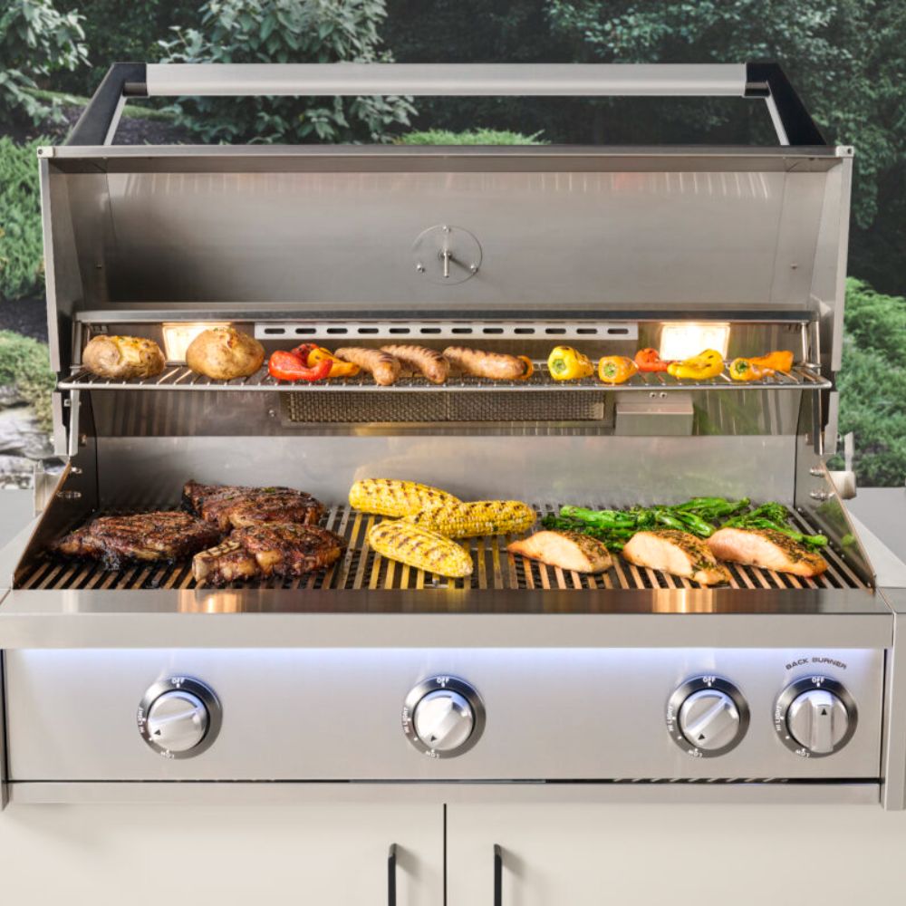 Stainless gas grill with open hood, grates filled with burgers, vegetables, and bread