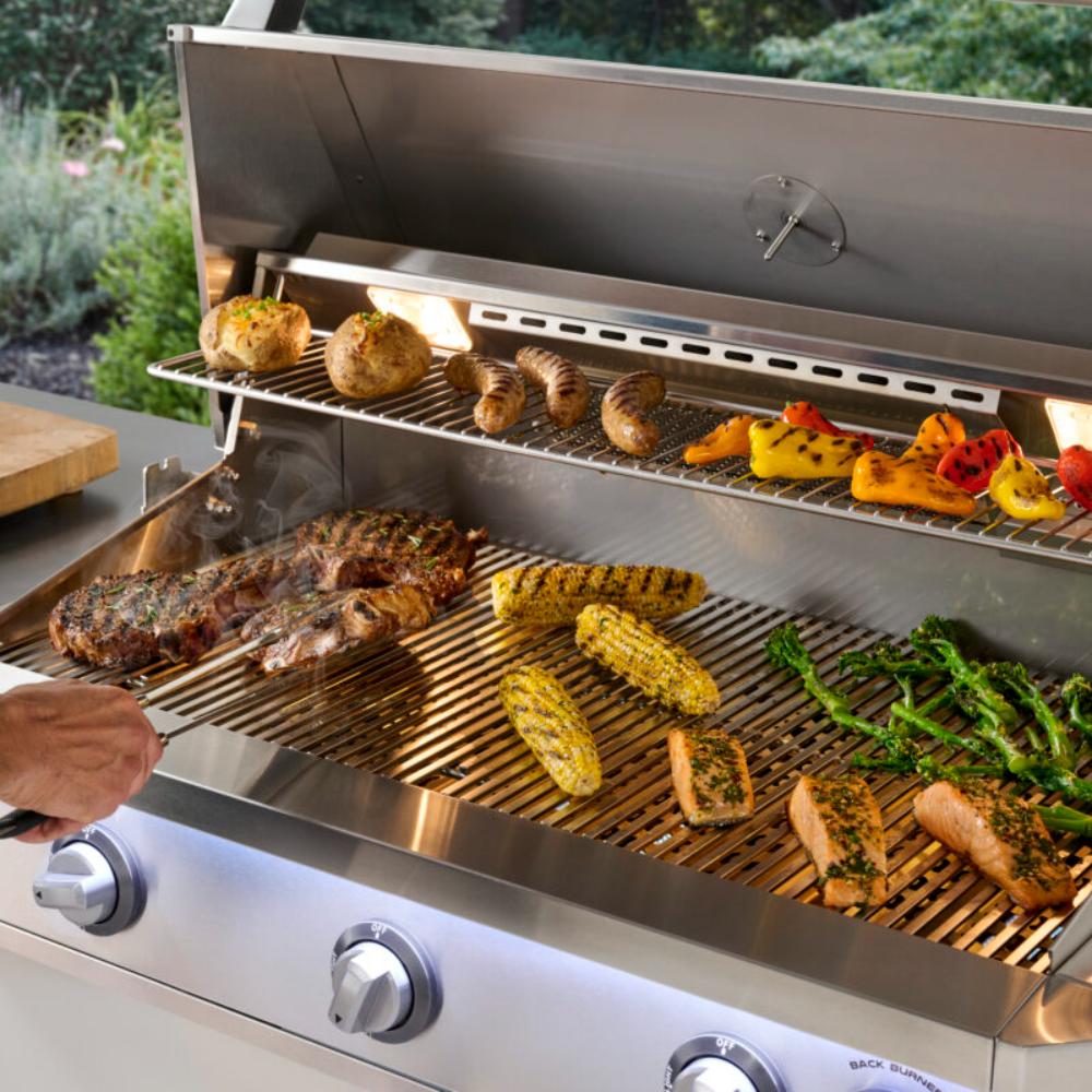 Stainless gas grill with open lid and food on grates, including corn and mixed vegetables