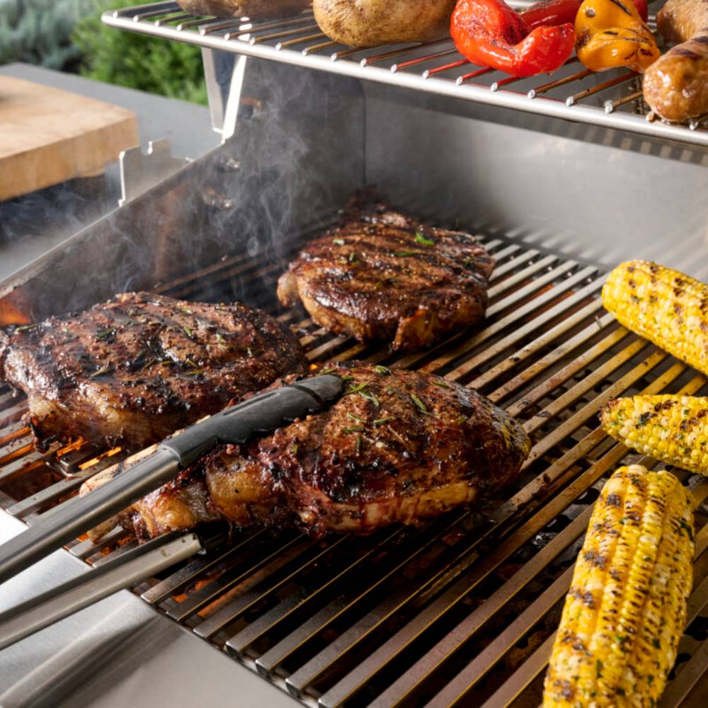 Open grill with searing meats and corn on black grates, stainless cooking surface