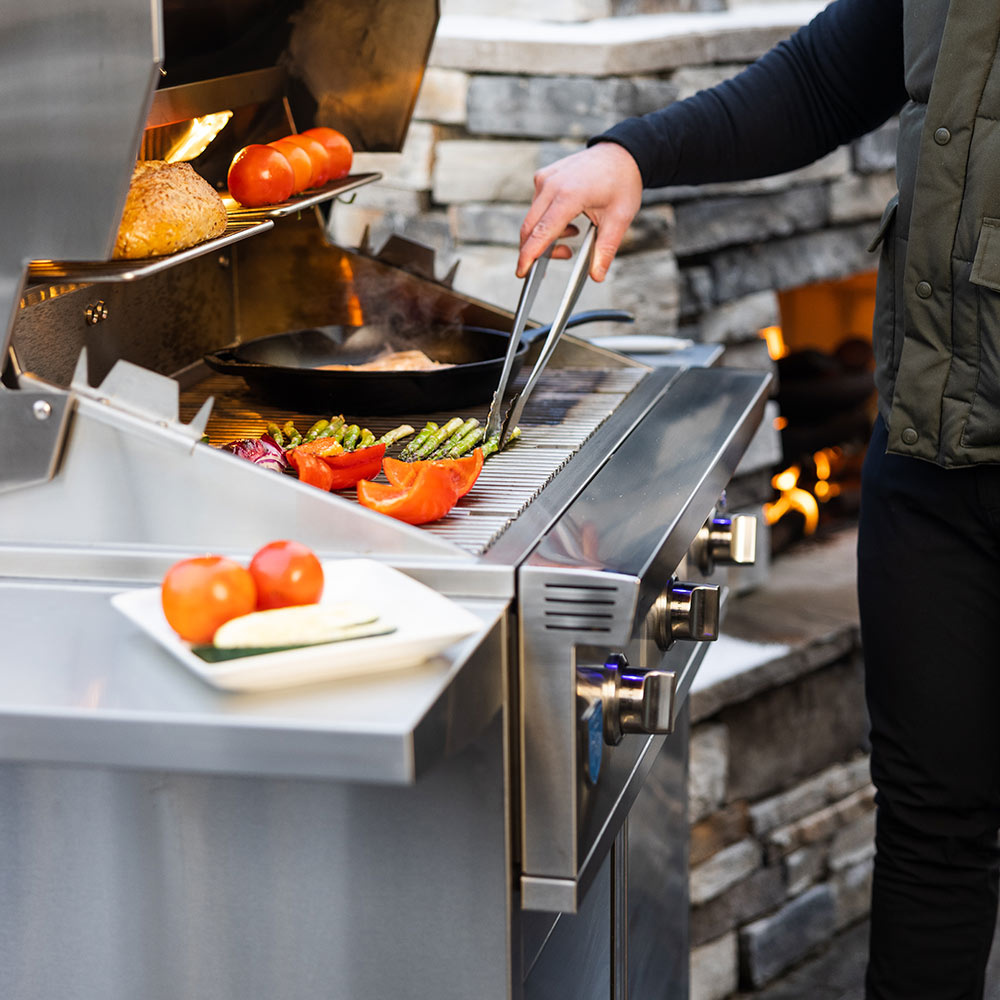 Stainless steel gas grill cooking with vegetables and tongs in the open lid