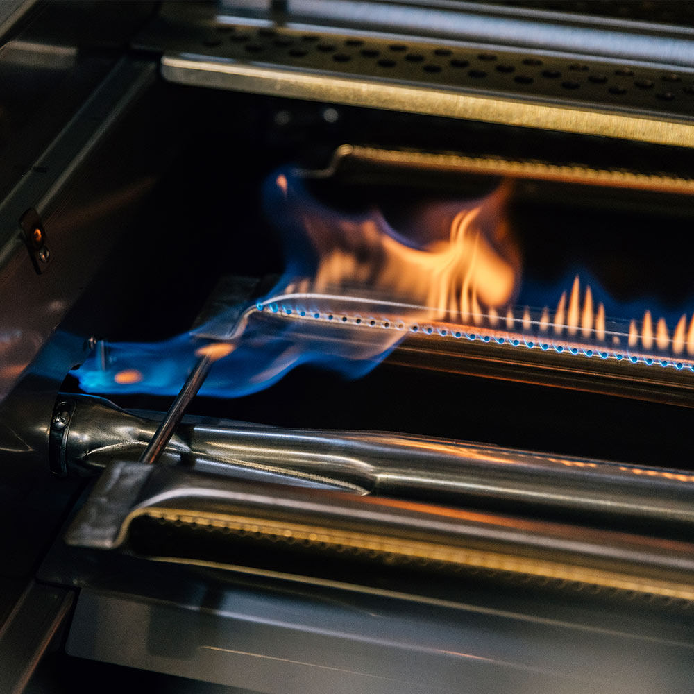 Gas grill burner area with orange flames and open firebox, front corner view