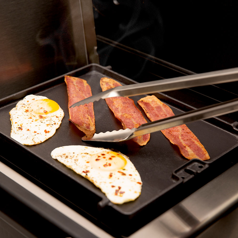 Cast iron side burner griddle with bacon, eggs, and tongs, steam visible