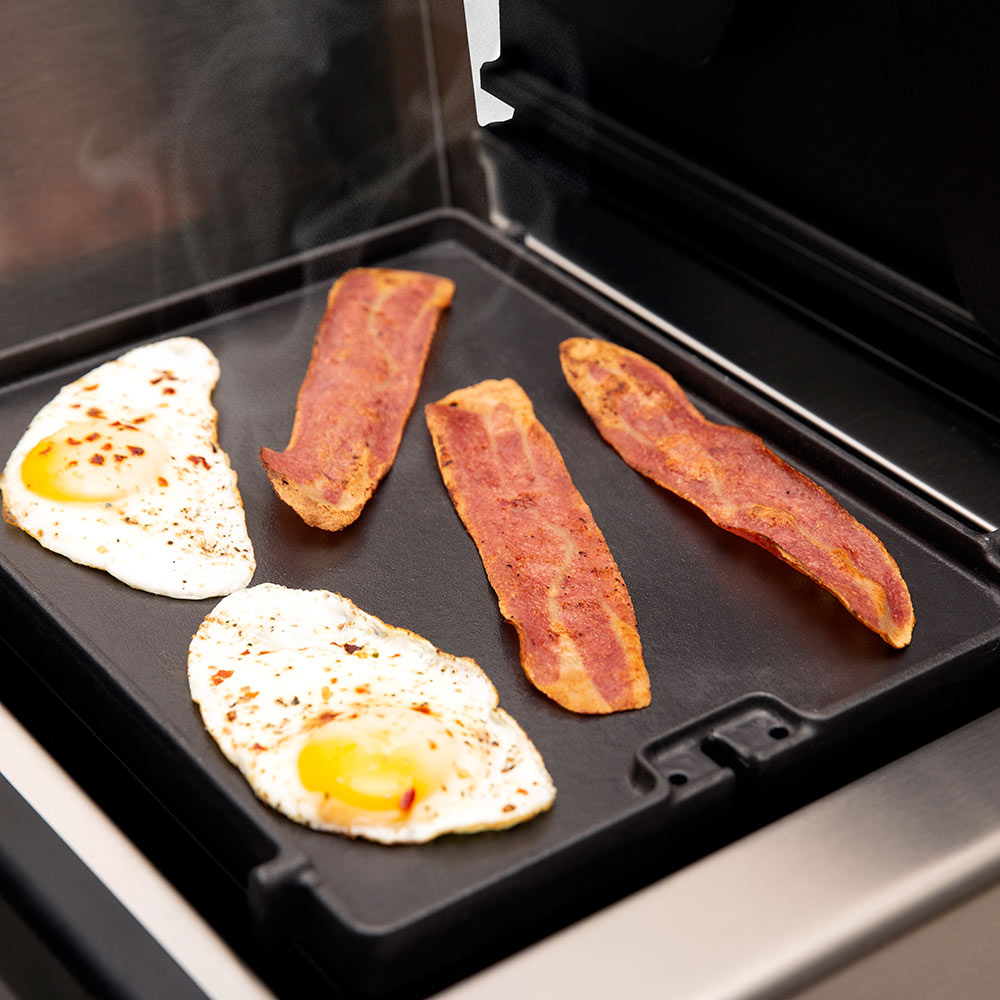 Cast iron side burner griddle with bacon and two fried eggs, steam rising