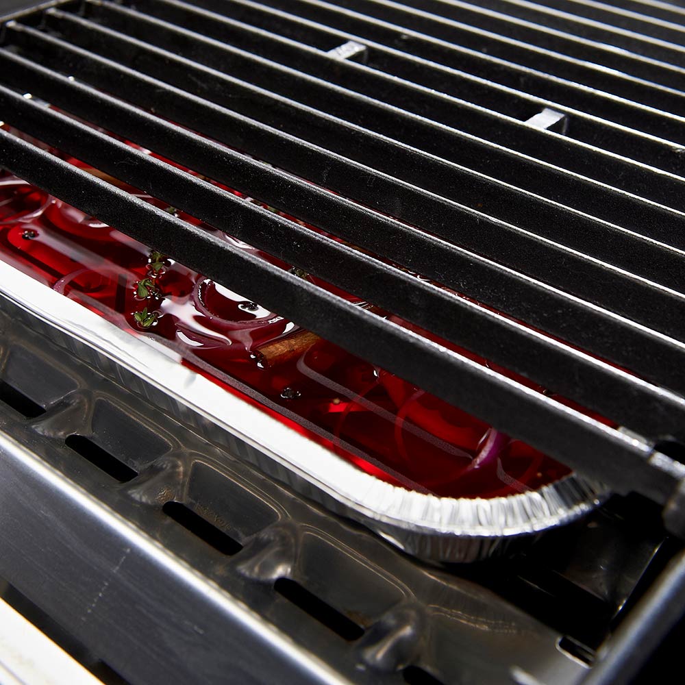 Aluminum drip pan on grill grates with red drippings and foil tray edge