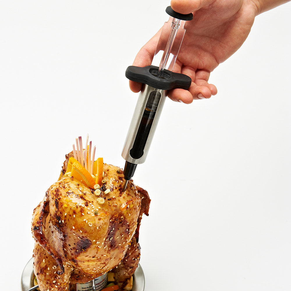 Marinade injector held by hand above roasted chicken; clear barrel and black grip