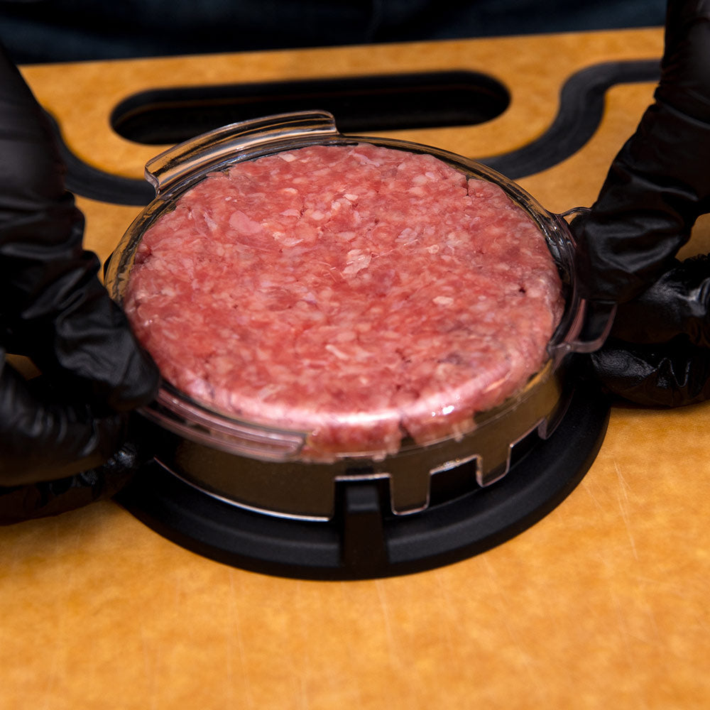 Raw burger patty under clear pressing lid on black base