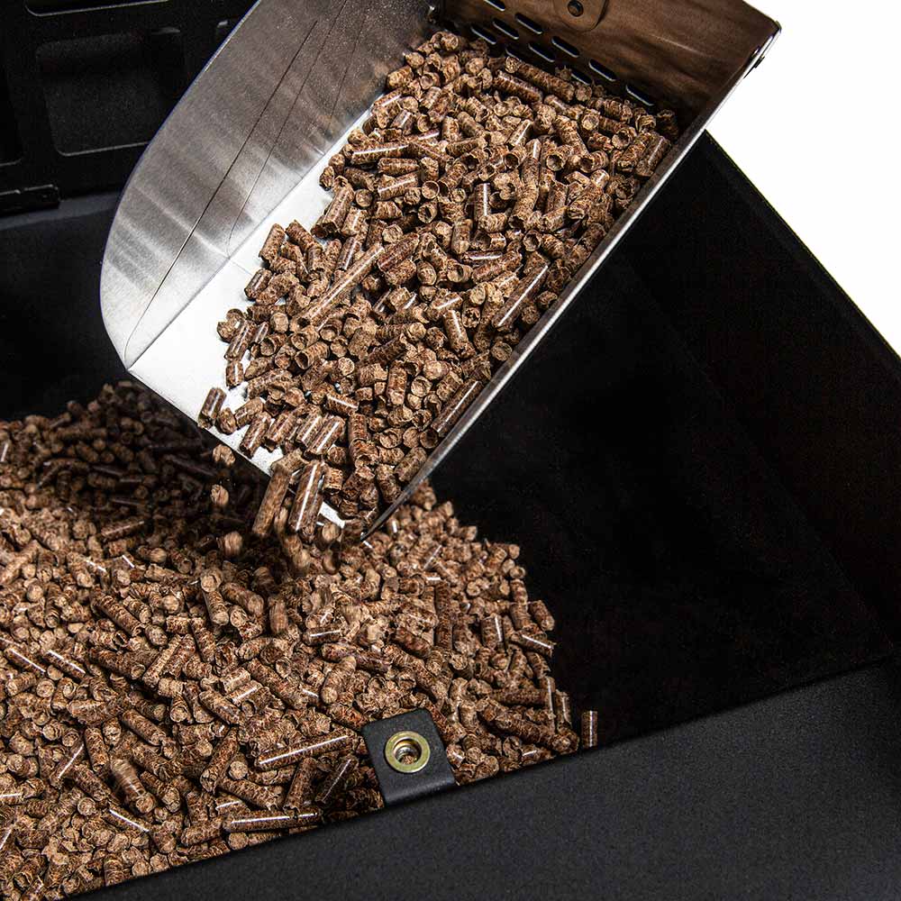 Tipped metal scoop dumping dark pellets into a grill tray