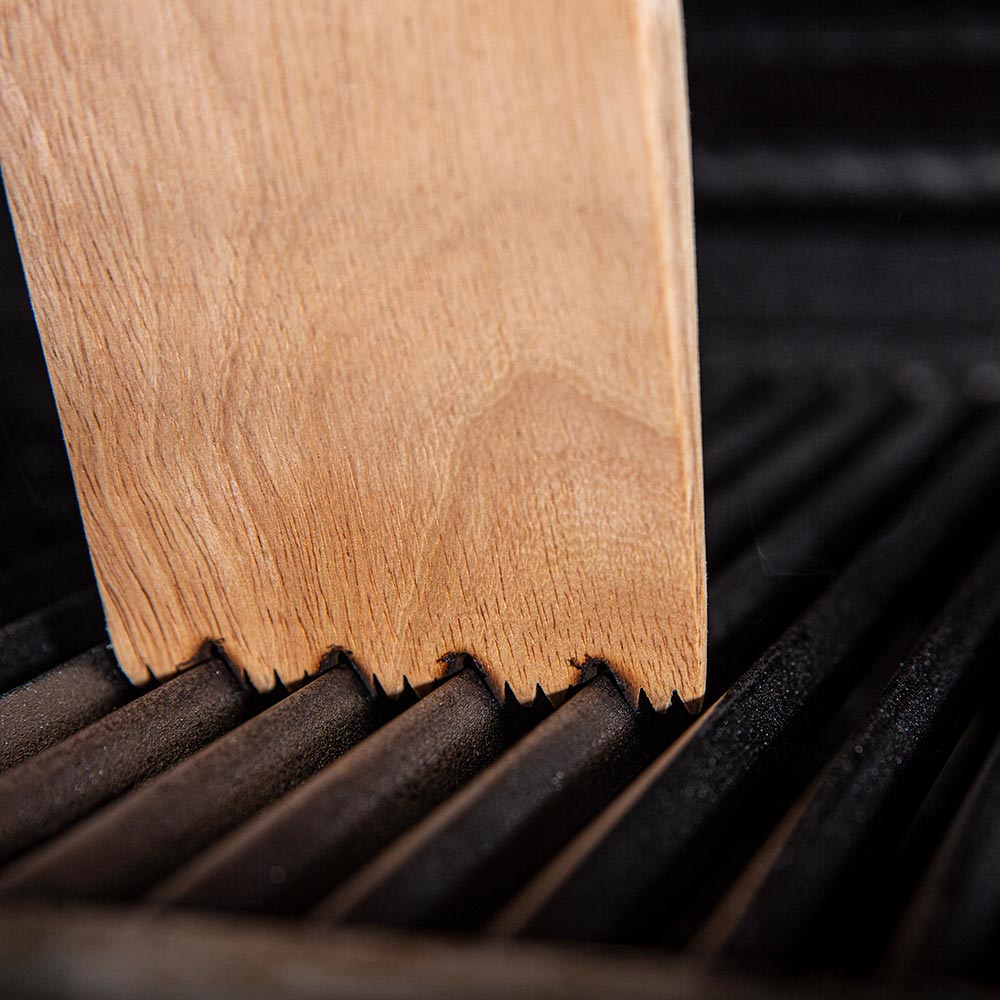 Wood BBQ grill scraper edge raking between grill grates, bristles visible