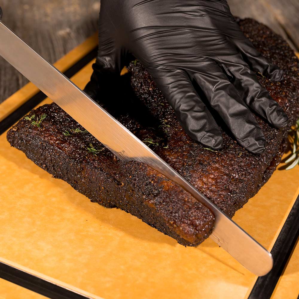 Wood fiber cutting board with two grilled meat logs being sliced by a gloved hand