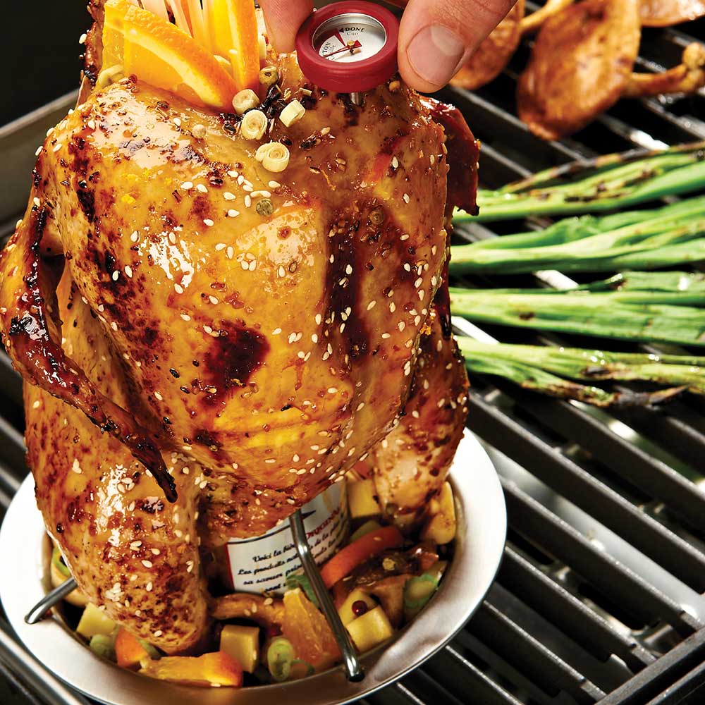 Roasted chicken on vertical roaster tray on a grill with vegetables and herbs