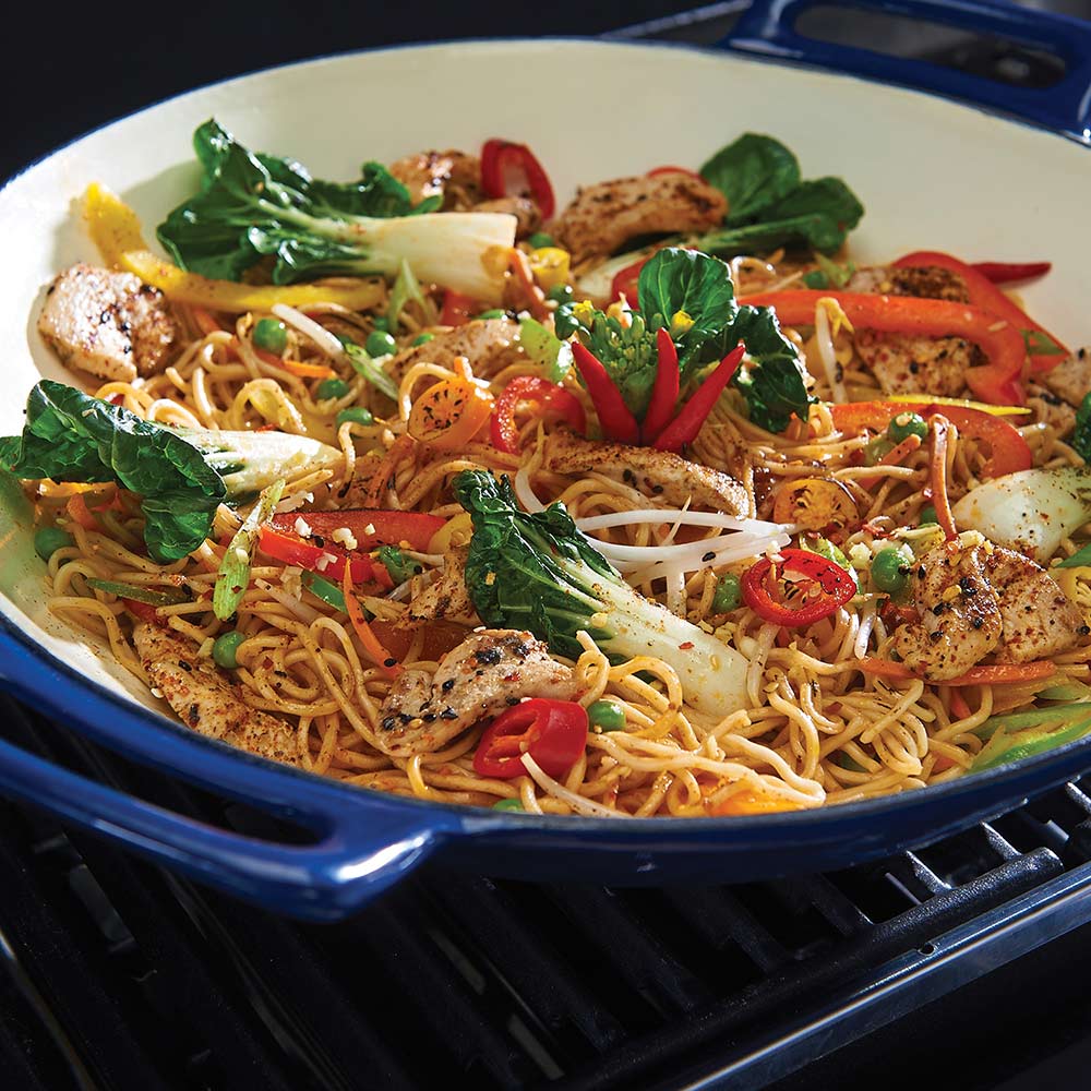 Porcelain cast iron wok on grill, close-up of noodle stir-fry with peppers and greens