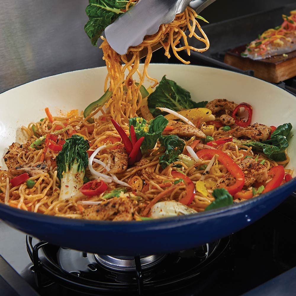 Porcelain cast iron wok on grill with noodle stir-fry being lifted with tongs