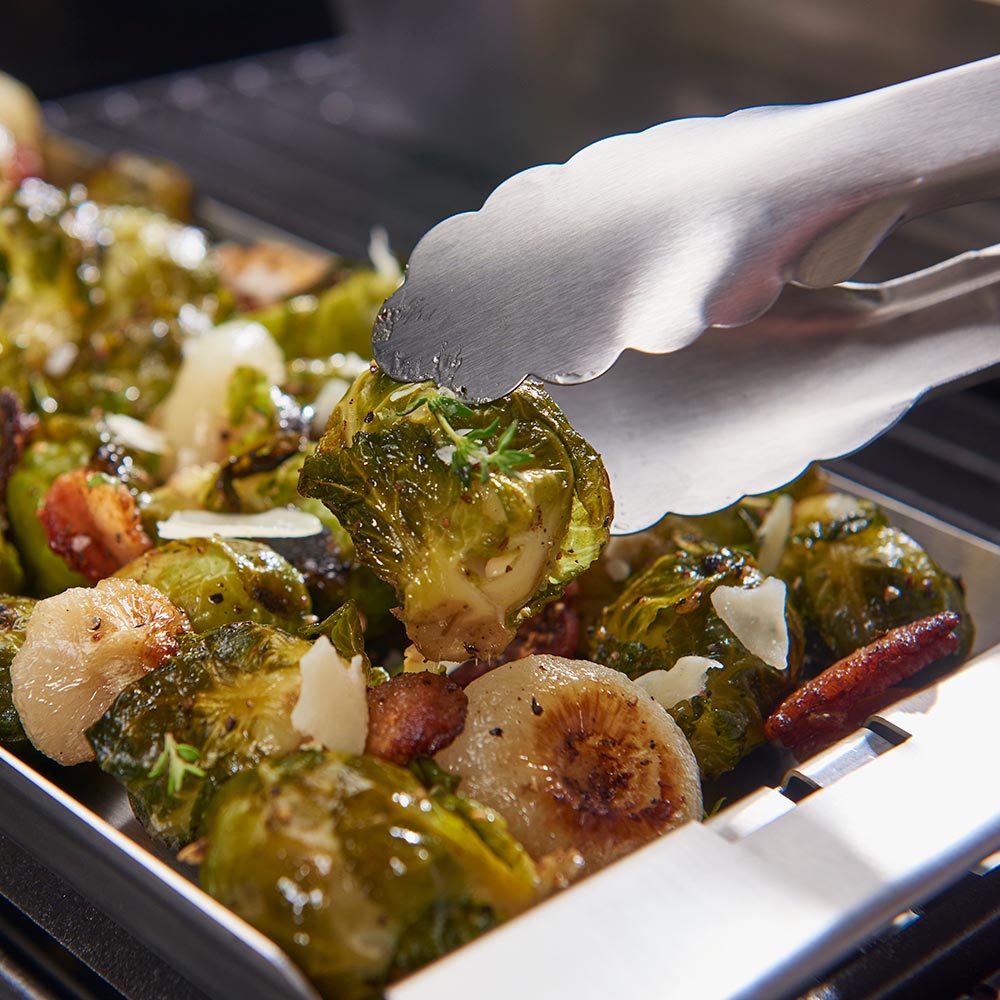 Gloved hand adding Brussels sprouts to a stainless steel grill topper tray