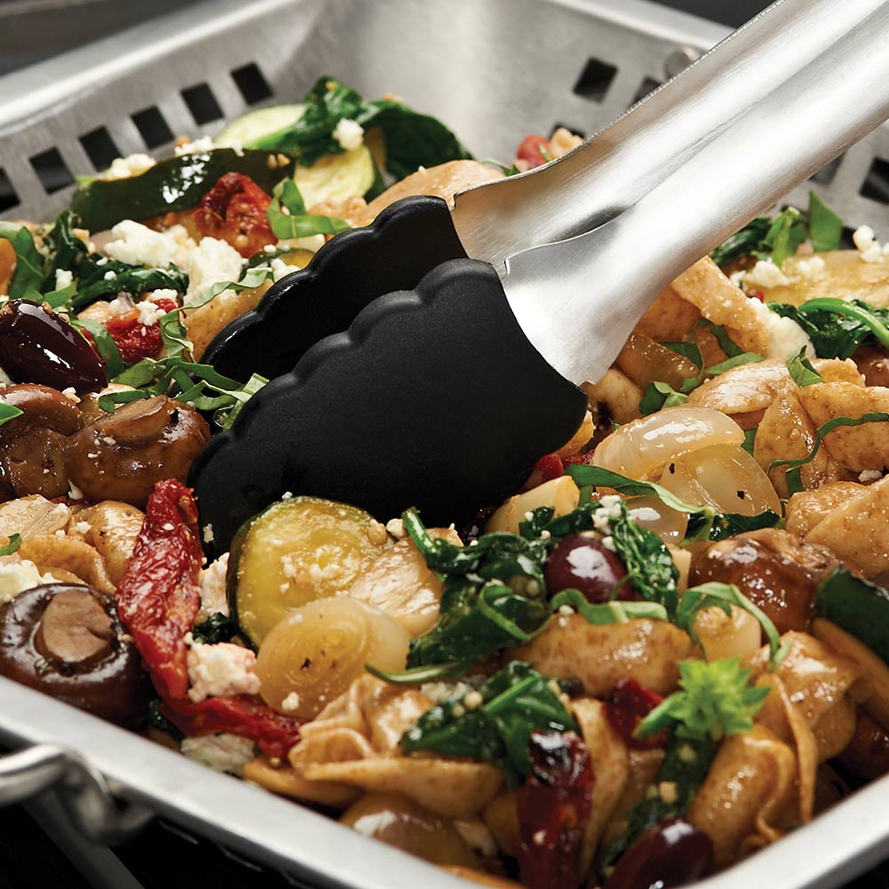 Stainless steel wok grill topper with side handles, holding grilled food and herbs