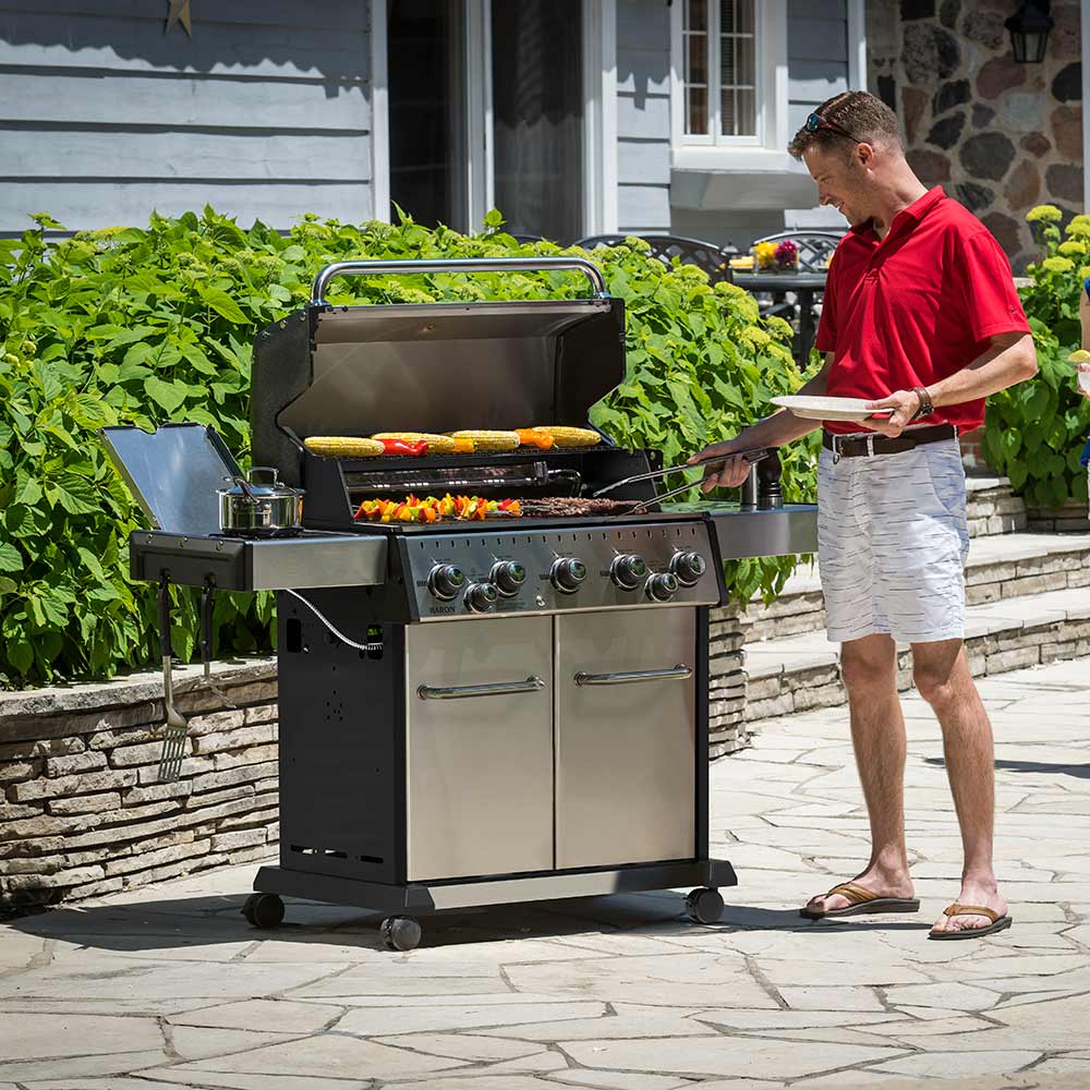 Stainless steel gas grill with rotisserie food and open grill lid beside side shelf