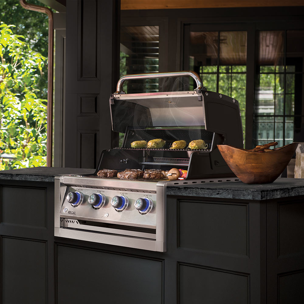 Built-in gas grill head with open lid and food cooking on grates, outdoor setting