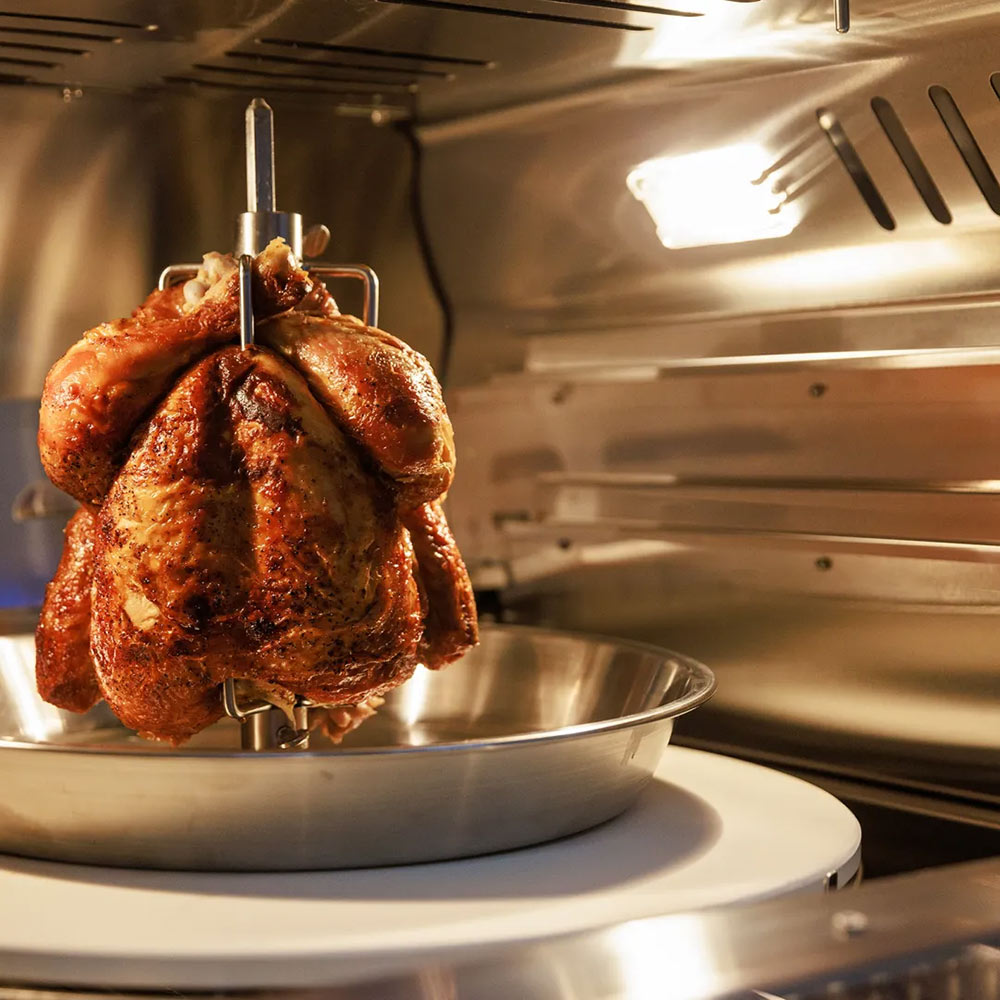 Rotisserie chicken on metal spit inside lit oven chamber with stainless interior