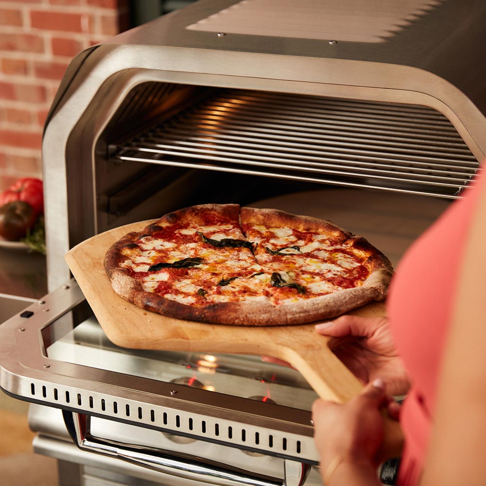 Wood peel placing pizza in open stainless oven chamber with lit burners