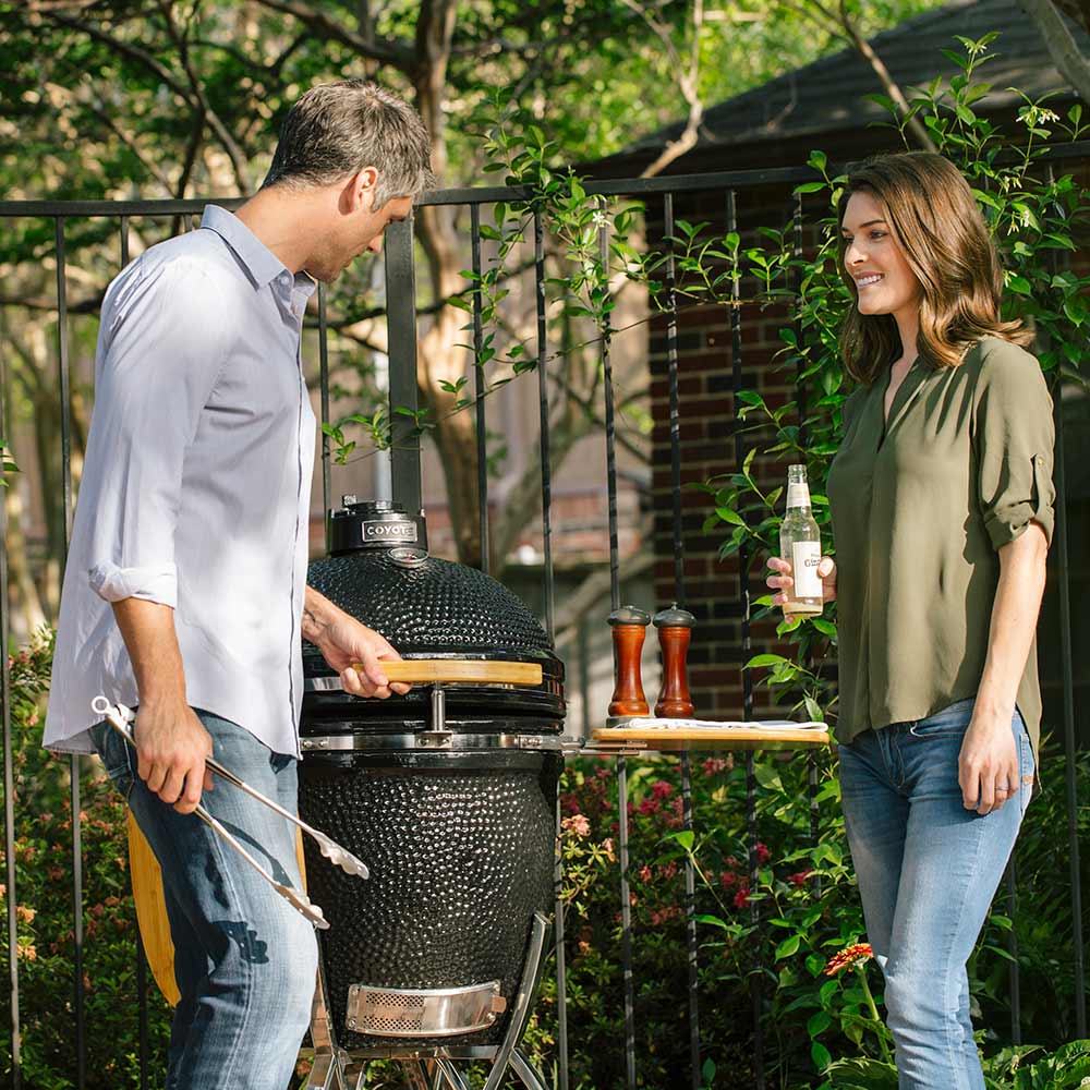 Ceramic smoker grill with open lid and wooden side shelves on metal wheeled stand
