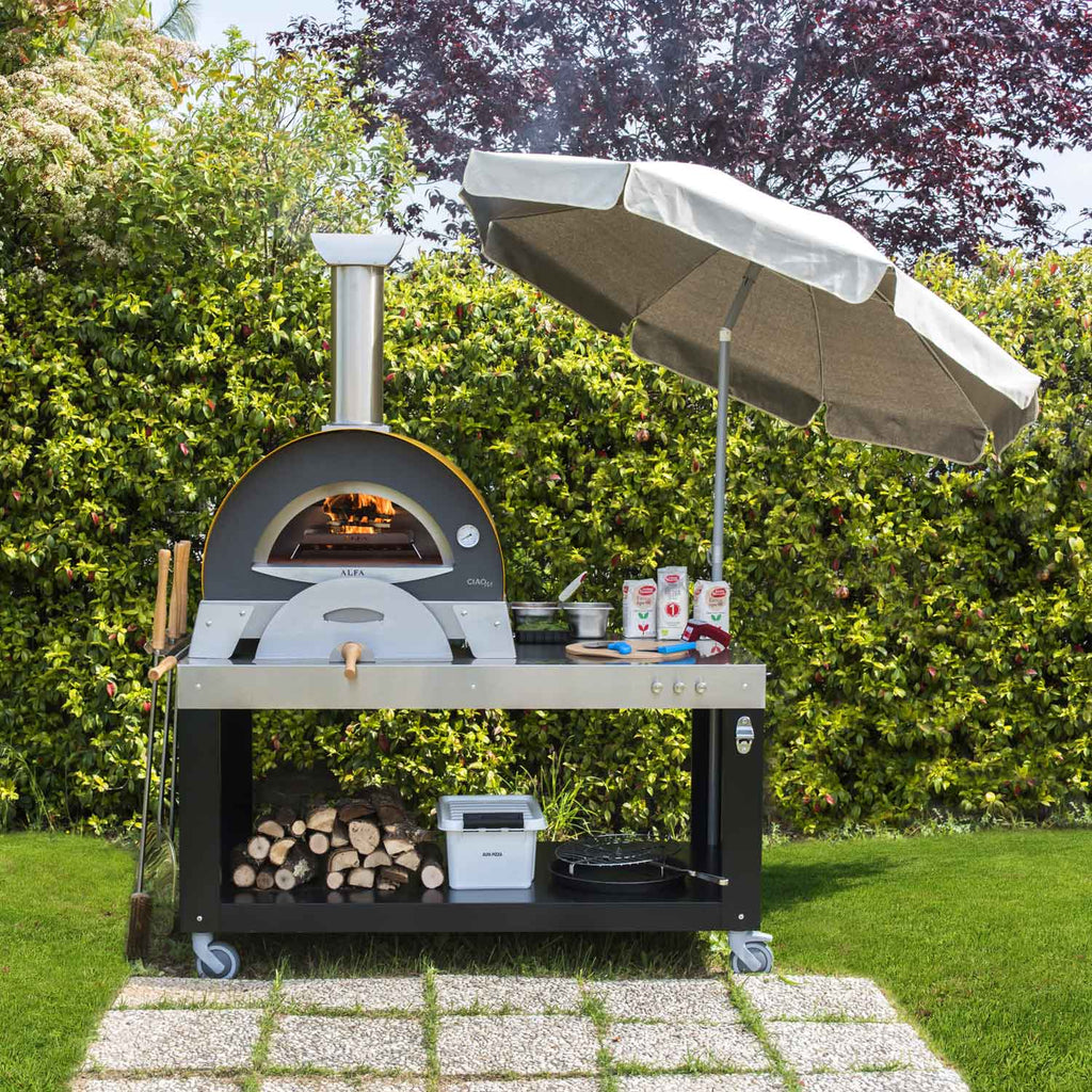 Countertop wood-fired pizza oven with black cooking dome and open flame, outdoor setup
