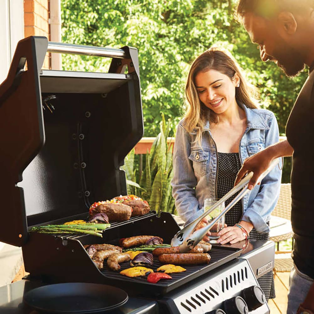 Gas grill on cart with open lid and burgers, vegetables, and a cooked tray