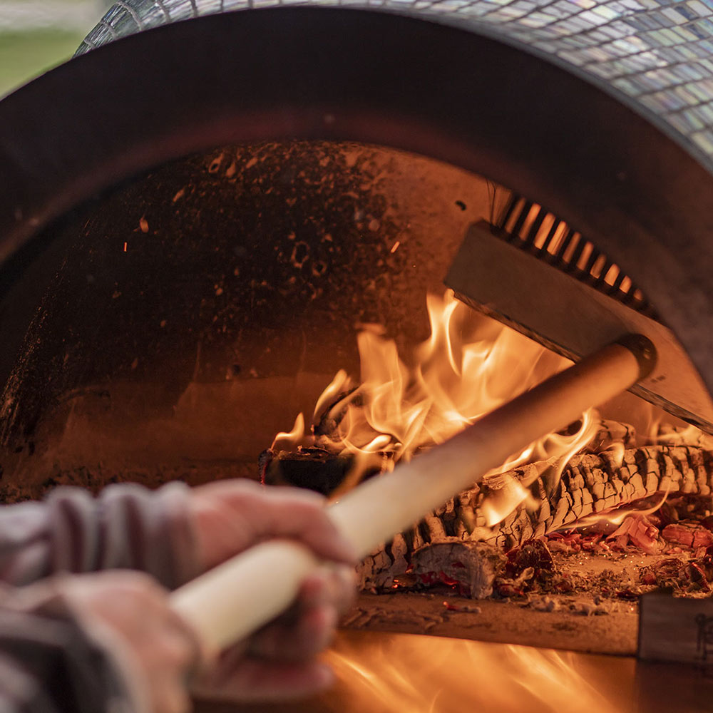 Pizza oven brush scrubbing the hot oven floor beside flames.