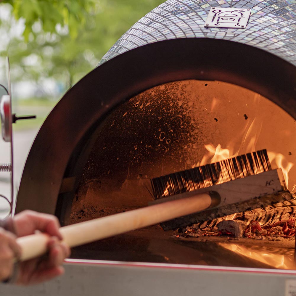 Pizza oven brush with wooden handle pushing ash in a burning oven.