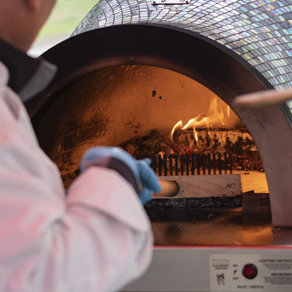 Pizza oven brush in gloved hand at the oven opening, flames burning.