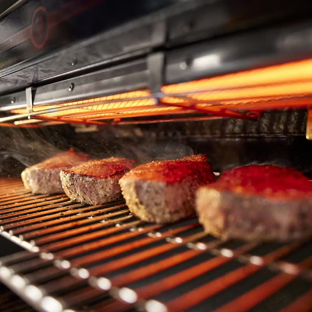 Steaks on grill grates with heat glow inside open gas grill, smoke visible