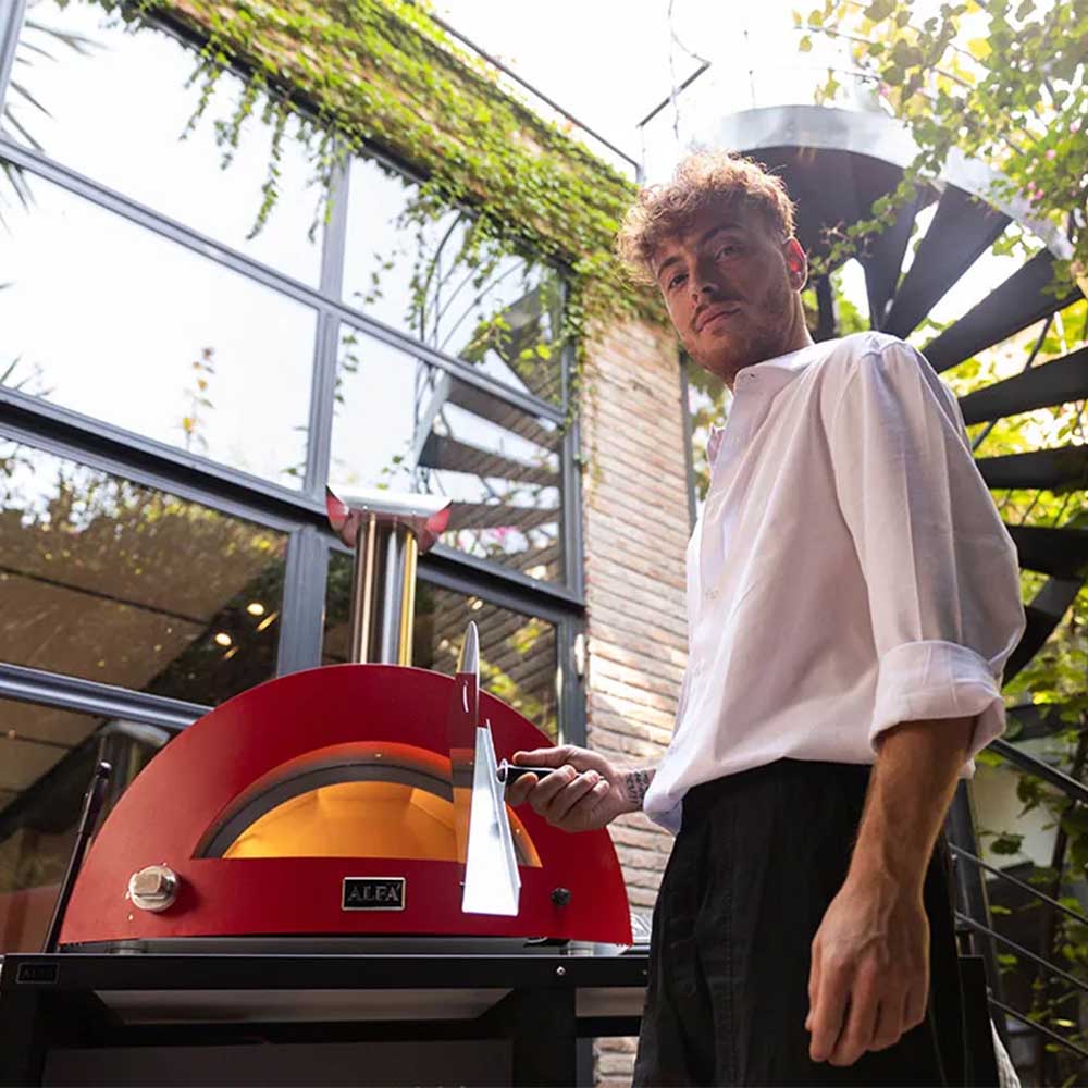 Red wood-fired pizza oven with cook holding a metal pizza peel near the front opening