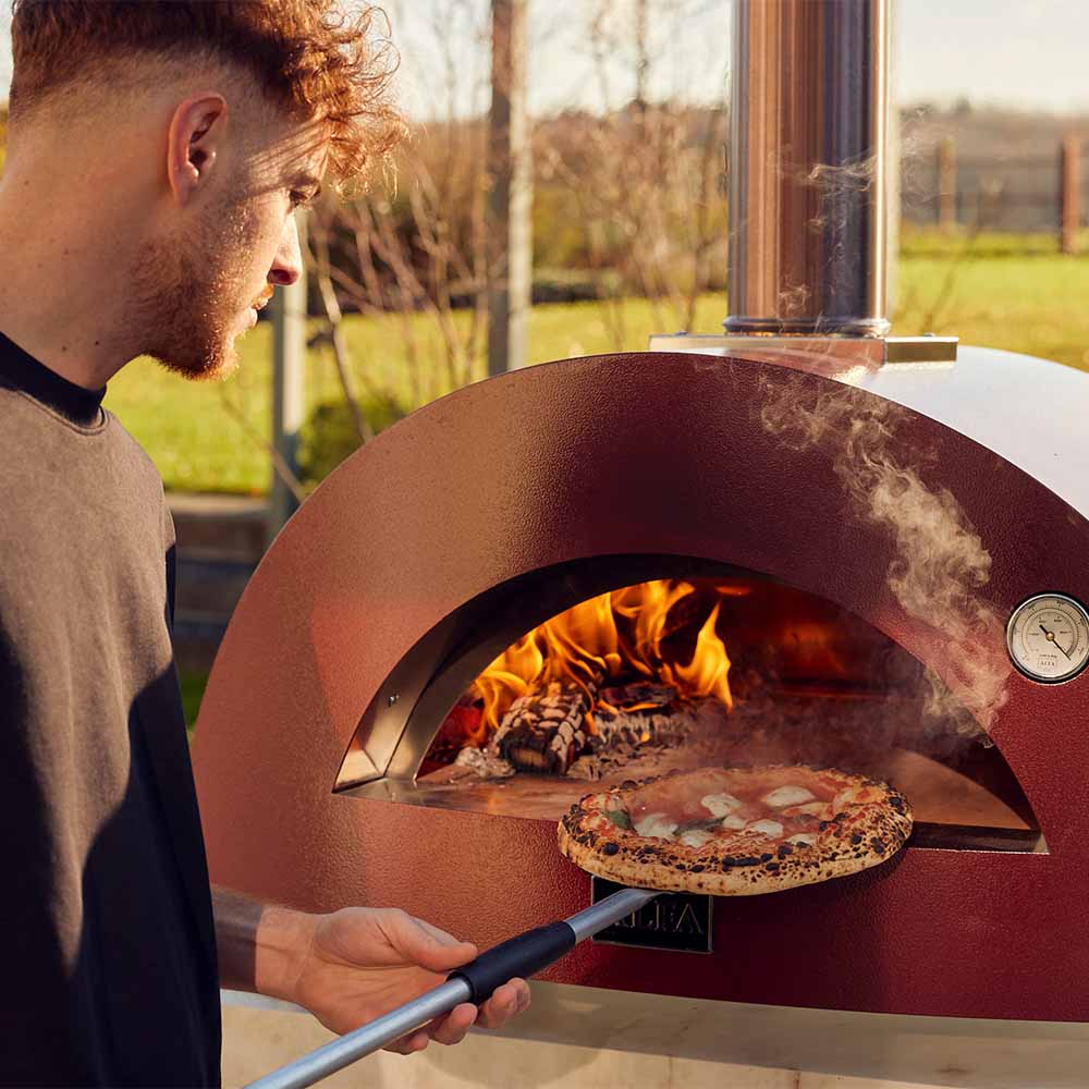 Antique red wood-fired pizza oven with pizza being placed on stone, flames inside