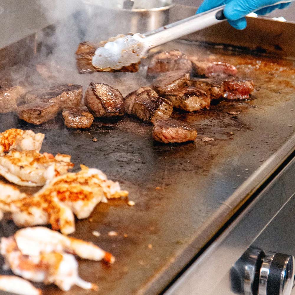 Flat-top gas griddle with sizzling meat and smoke, flame tamer surfaces visible