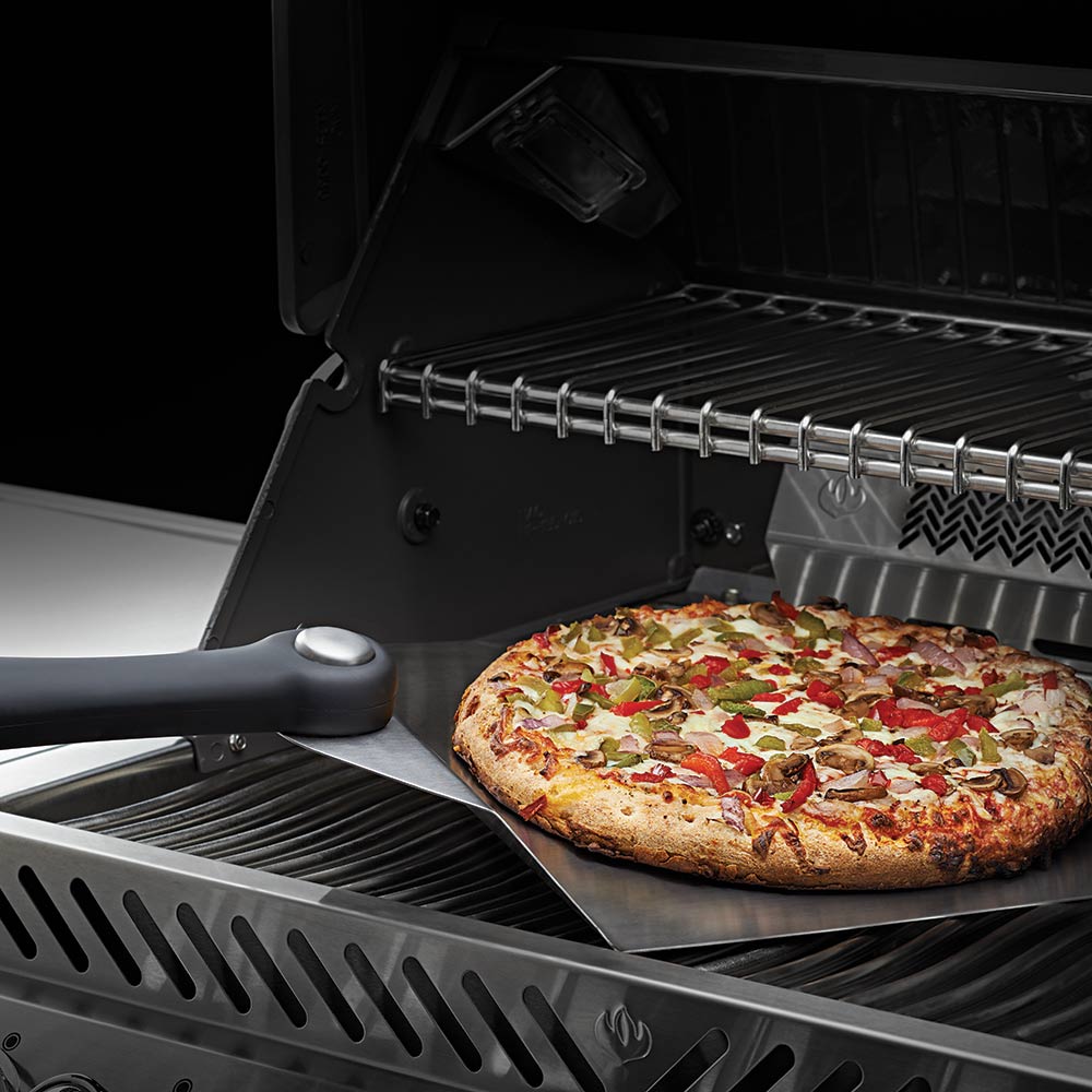 Pizza spatula lifting a large pizza from a grill grate with black handle