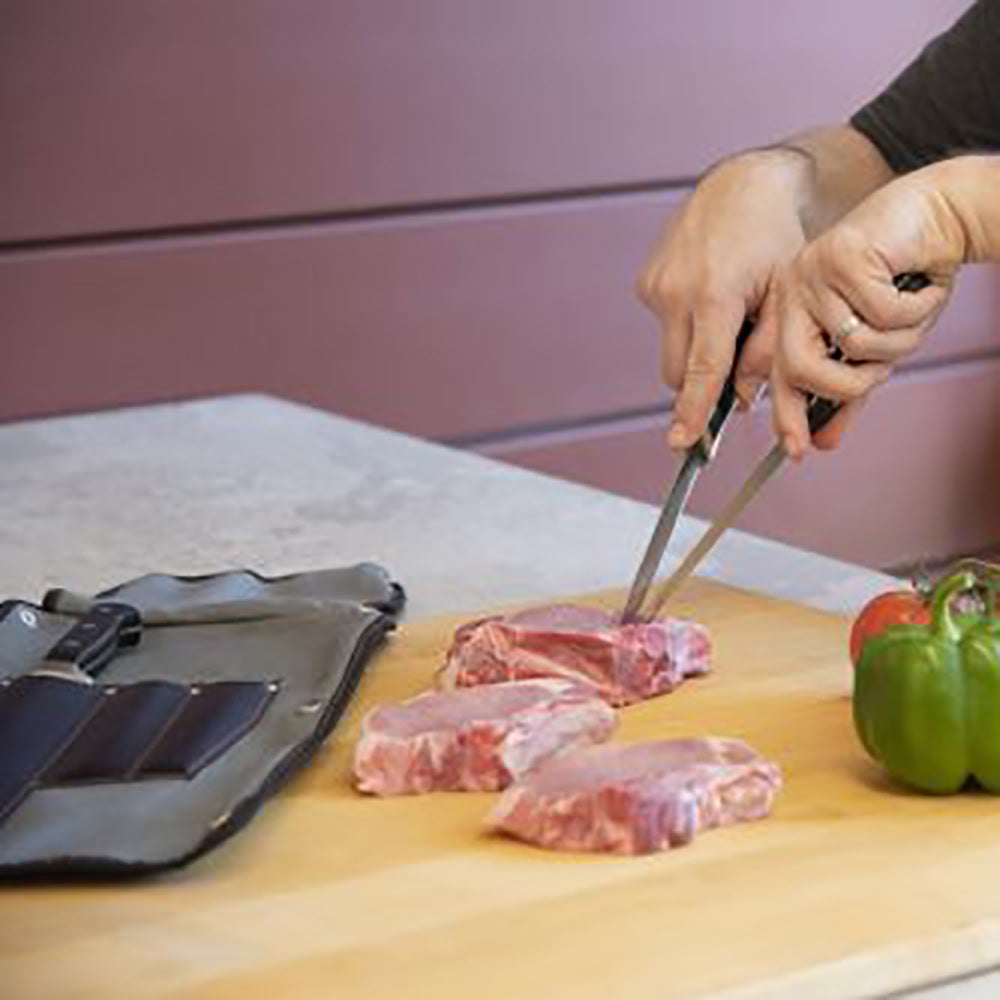 Butcher knife cutting meat on a wooden board next to an open knife roll