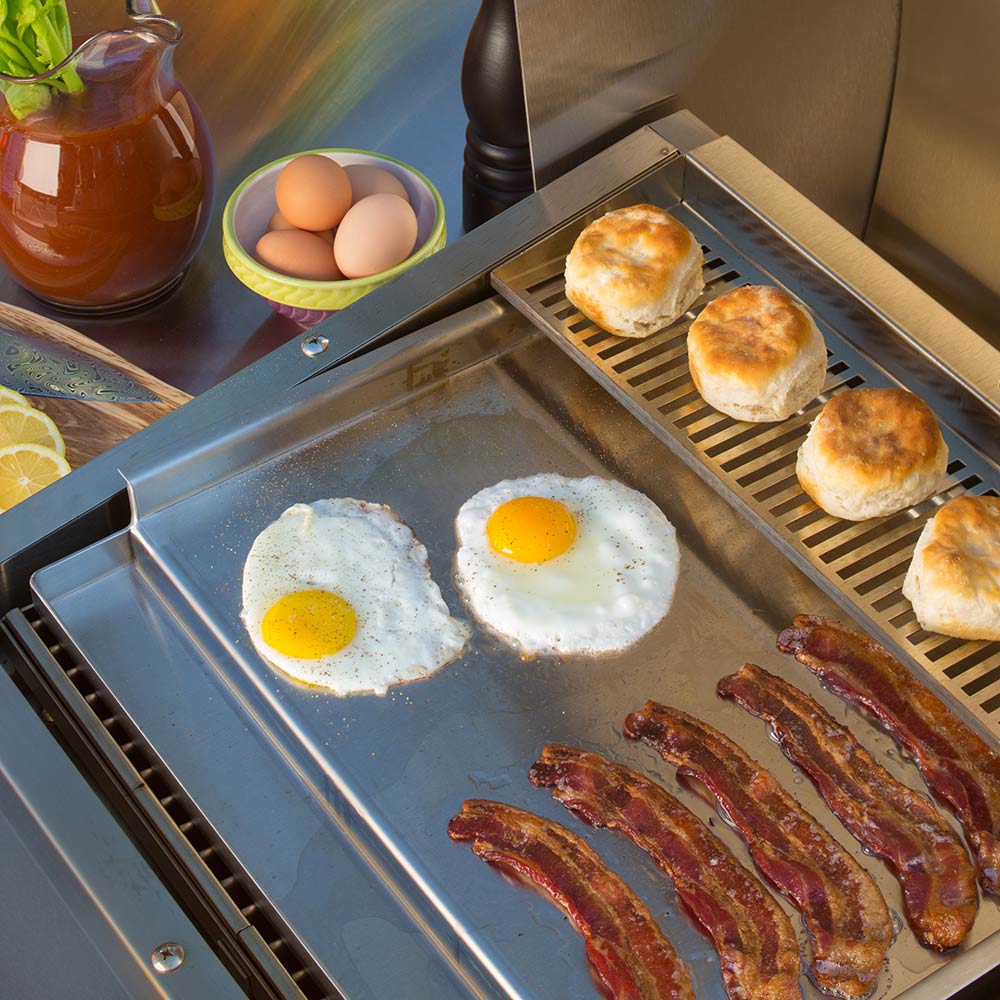 Stainless steel griddle with burgers, sunny-side eggs, and bacon strips
