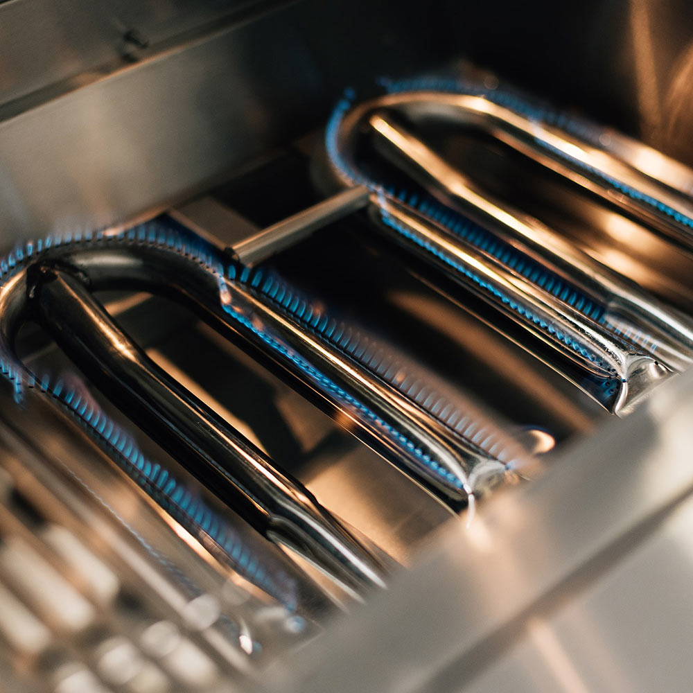 Gas grill burner tubes inside the firebox with glowing flames under the cooking surface