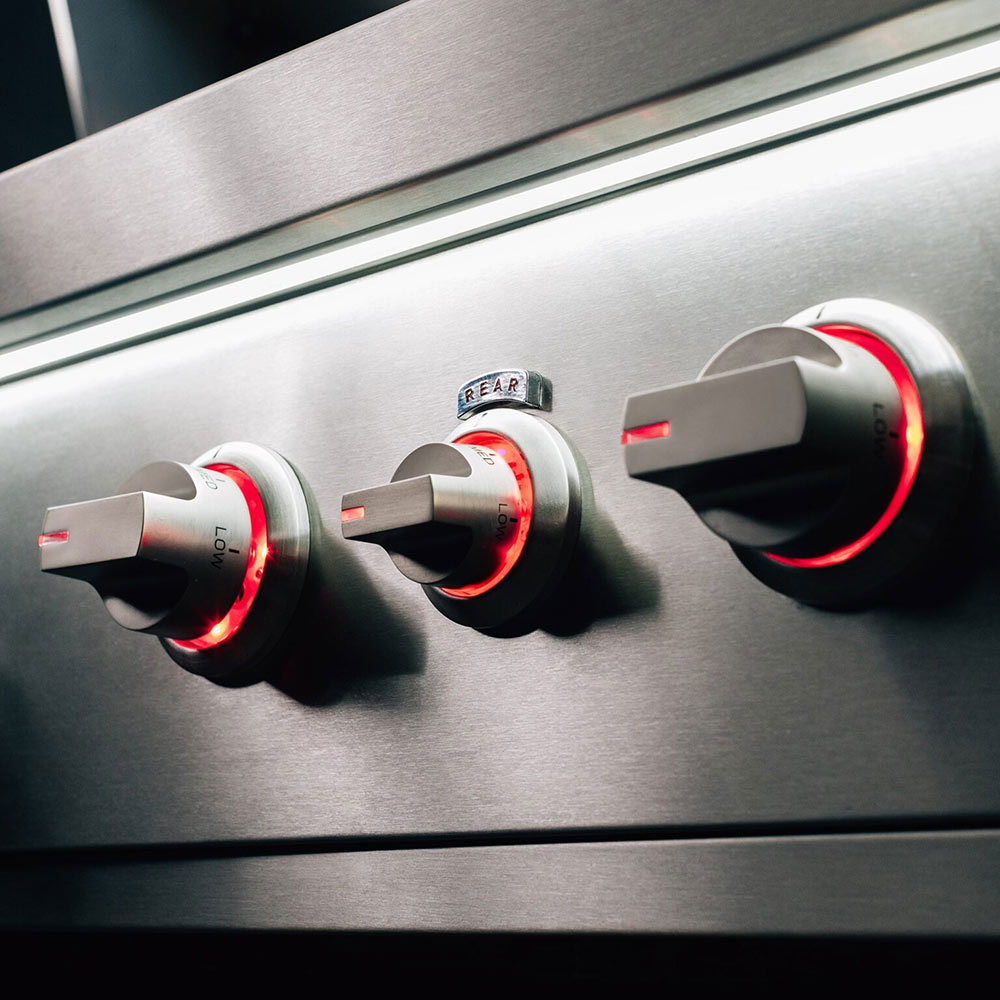 Gas grill control panel with four stainless knobs and red indicator rings.