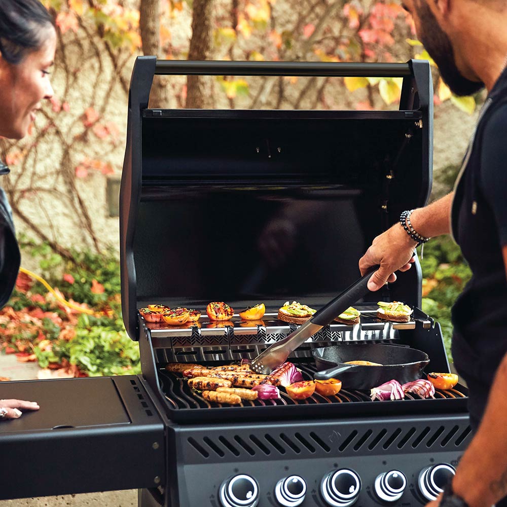 Open black gas grill with food cooking on grates and in side trays