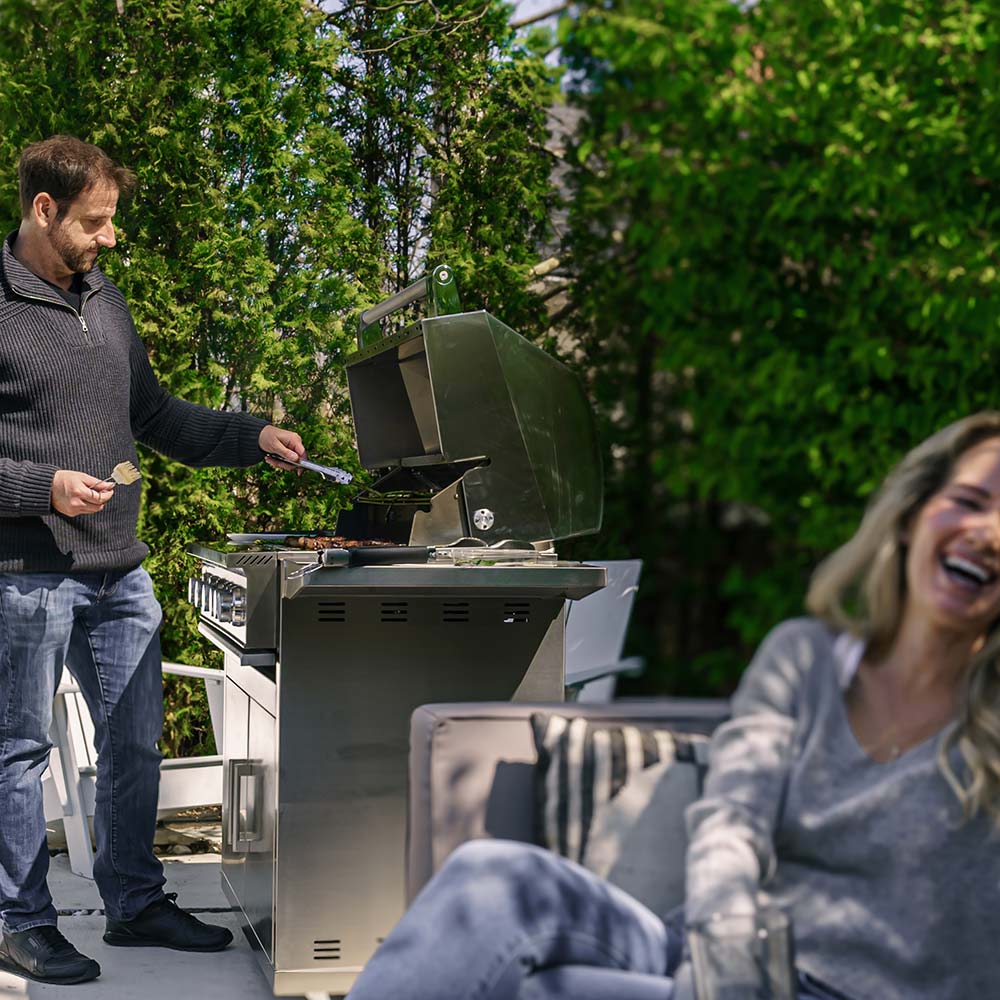 Outdoor gas grill on cart with open hood as someone tends the cooking area